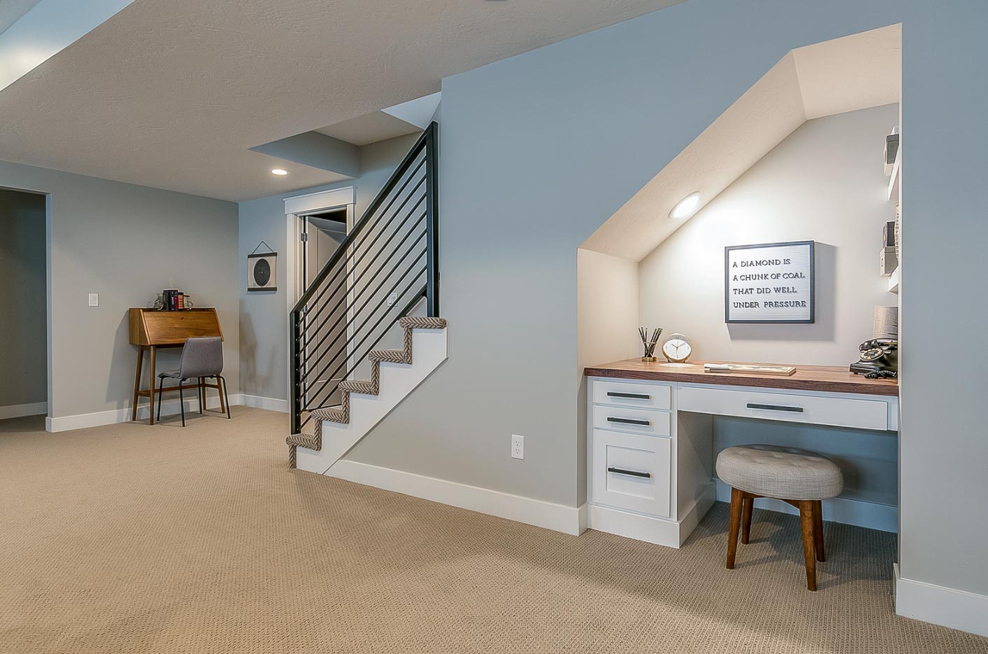 Cozy home office nook under stairs with desk, chair, and motivational sign, expertly crafted by MD's finest home builders. Nearby, a small reading area with a lamp invites relaxation.