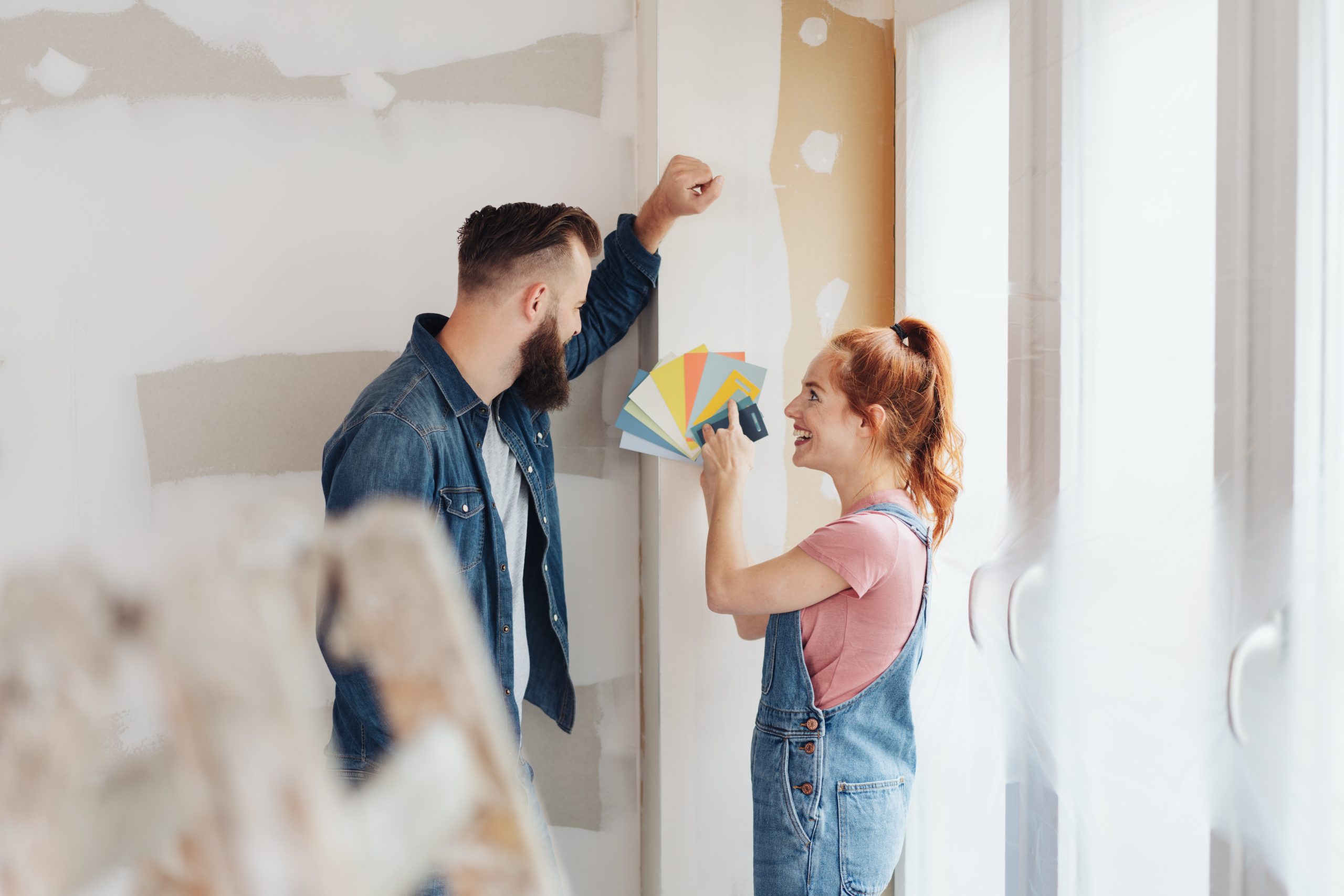 A couple discusses paint colors, holding swatches while renovating a room with unfinished walls.