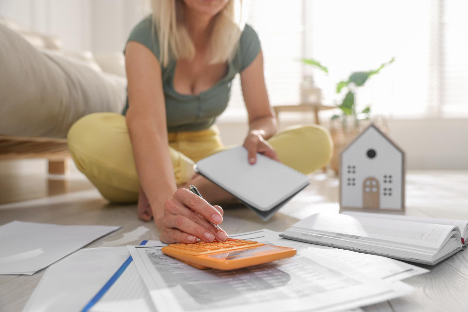103484 Woman using calculator on floor with papers, notepad, and model house, planning home finances.